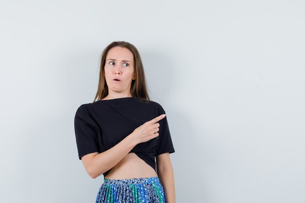 Junge Frau, die rechts mit Zeigefinger im schwarzen T-Shirt und im blauen Rock zeigt und überrascht schaut