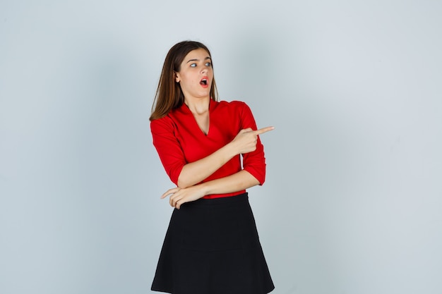 Junge Frau, die rechts mit Zeigefinger in roter Bluse, schwarzem Rock zeigt und überrascht schaut
