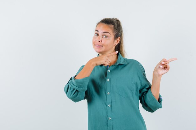 Junge Frau, die rechts mit Zeigefingern in grüner Bluse zeigt und niedlich schaut