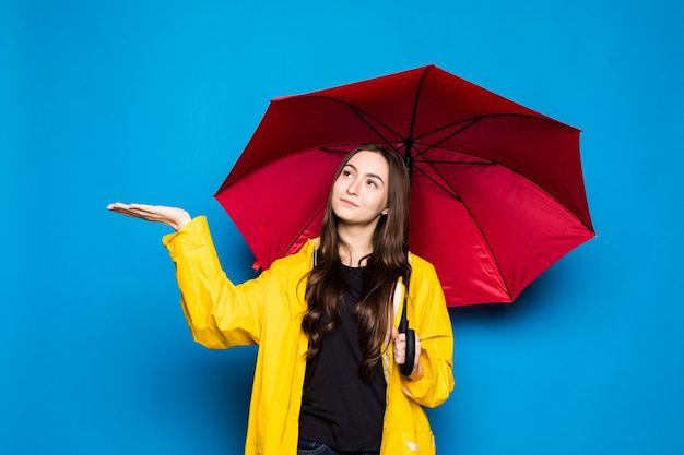 Junge Frau, die Regenmantel hält, der bunten Regenschirm über blauer Wand hält