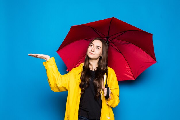 Junge Frau, die Regenmantel hält, der bunten Regenschirm über blauer Wand hält