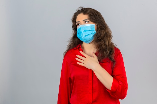 Junge Frau, die rote Bluse in der medizinischen Schutzmaske trägt, die unwohl und krank mit Hand auf Brust über lokalisiertem weißem Hintergrund schaut