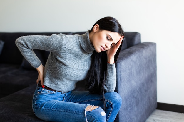 Junge Frau, die Rückenschmerzen leidet und sich beschwert, auf einer Couch im Wohnzimmer zu Hause zu sitzen