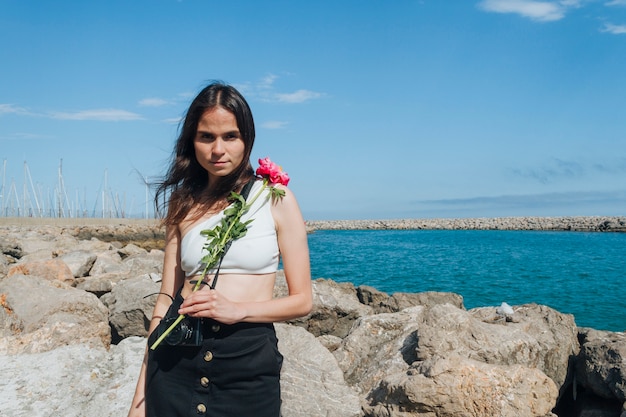 Junge Frau, die schöne Blume hält und die Kamera steht nahes Meer betrachtet