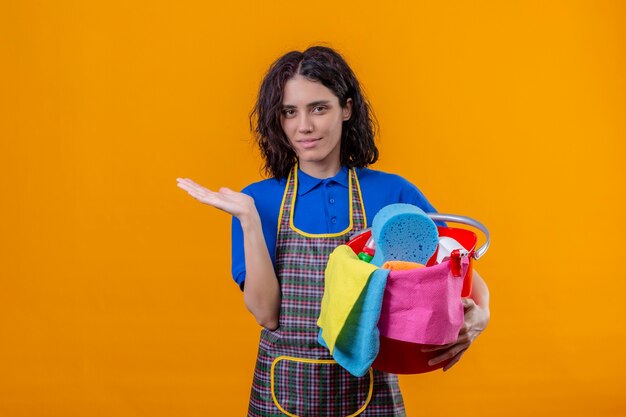 Junge Frau, die Schürze hält Eimer mit Reinigungswerkzeugen präsentiert, die mit Arm der Hand präsentieren, die zuversichtlich steht über orange Hintergrund