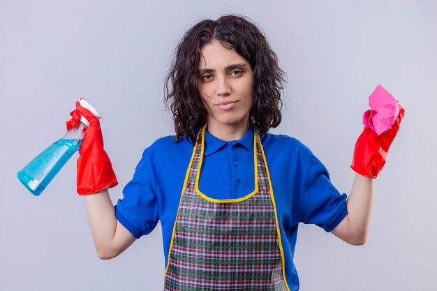 Junge Frau, die Schürze und Gummihandschuhe trägt, die Reinigungsspray und Teppich halten, die Hände mit skeptischem Ausdruck über weißer Wand ausbreiten