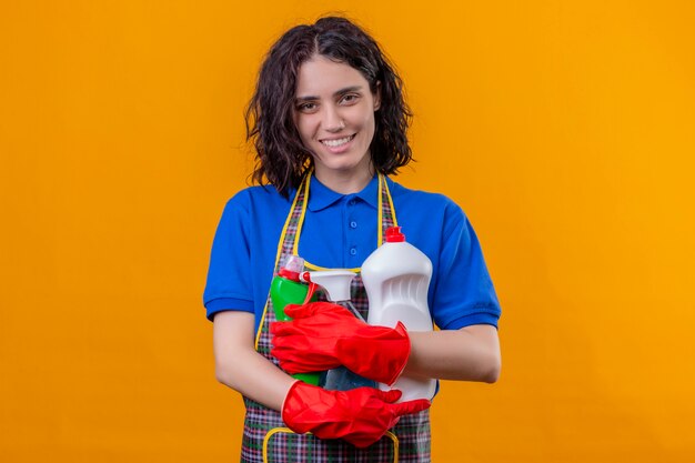 Junge Frau, die Schürze und Gummihandschuhe trägt, die Reinigungsvorräte positiv und glücklich lächelnd über orange Wand halten