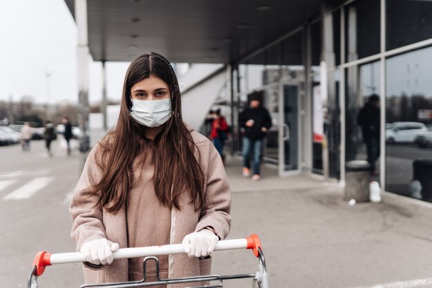 Junge Frau, die Schutzgesichtsmaske gegen Coronavirus 2019-nCoV trägt, der einen Einkaufswagen schiebt.