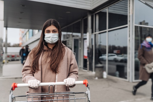 Junge Frau, die Schutzgesichtsmaske gegen Coronavirus trägt, der einen Einkaufswagen drückt.