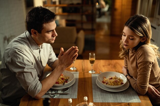 Junge Frau, die sich traurig fühlt, während ihr Freund versucht, sich beim Abendessen am Esstisch zu entschuldigen