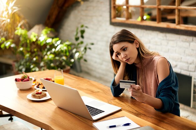 Junge Frau, die sich traurig fühlt, während sie zu Hause ihr Online-Bankkonto über den Laptop überprüft