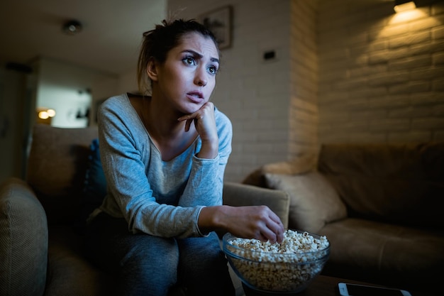Junge Frau, die sich überrascht fühlt, während sie Filme anschaut und spät in der Nacht zu Hause Popcorn isst