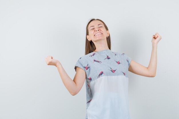 Junge Frau, die Siegergeste im T-Shirt zeigt und glückliche Vorderansicht schaut.