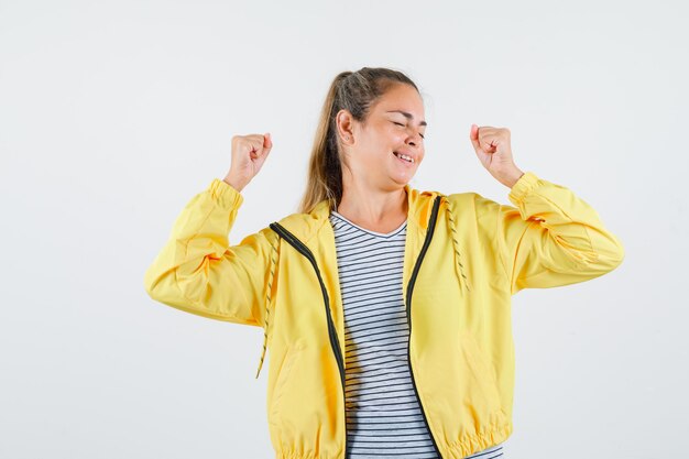 Junge Frau, die Siegergeste in Jacke, T-Shirt zeigt und glücklich schaut. Vorderansicht.