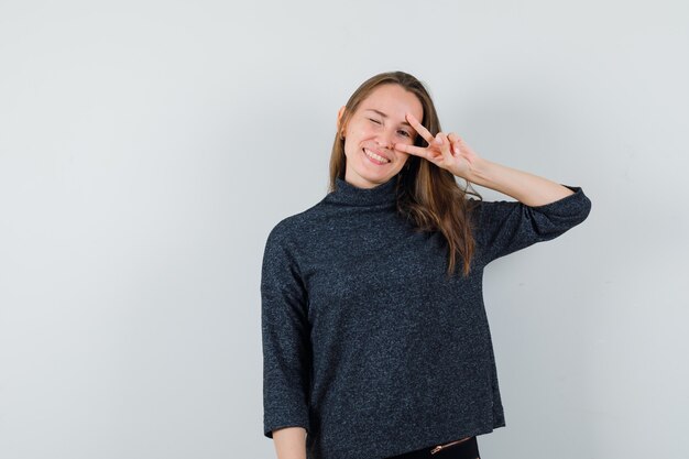 Junge Frau, die Siegesgeste im Hemd zeigt und zuversichtlich schaut. Vorderansicht.