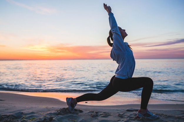 Junge Frau, die Sportübungen am Sonnenaufgangstrand am Morgen tut