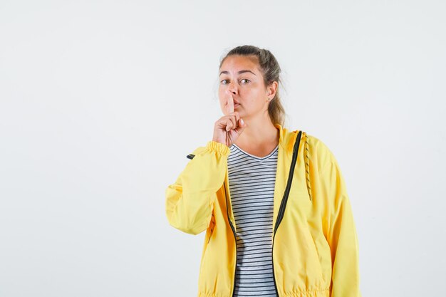 Junge Frau, die Stille Geste im T-Shirt, in der Jacke zeigt und vorsichtig schaut. Vorderansicht.