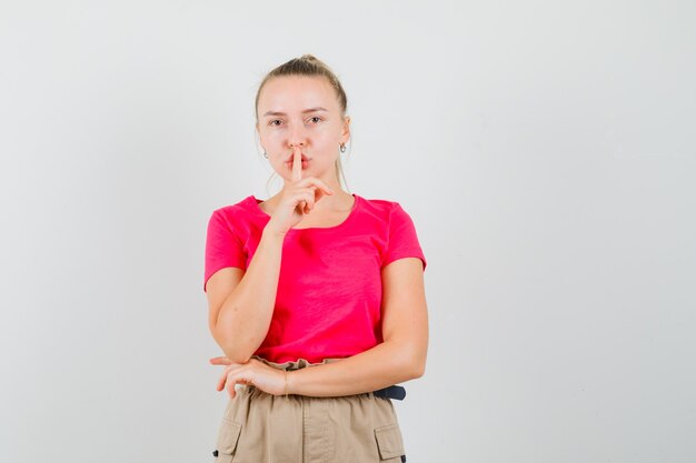 Junge Frau, die Stille Geste im T-Shirt und in der Hose zeigt und vorsichtig schaut