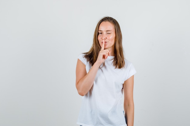 Junge Frau, die Stille Geste mit blinzelndem Auge in der Vorderansicht des weißen T-Shirts zeigt.