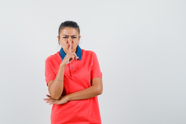 Junge Frau, die Stille Geste zeigt, Zeigefinger auf Mund in rotem T-Shirt setzt und wütend, Vorderansicht schaut.