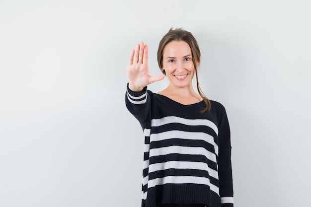 Junge Frau, die Stoppschild mit einer Hand in gestreiften Strickwaren und in schwarzen Hosen zeigt und glücklich schaut