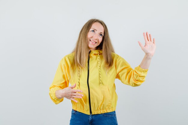 Junge Frau, die Stoppschild zeigt und in der gelben Bomberjacke und in der blauen Jeans lächelt und optimistisch schaut. Vorderansicht.