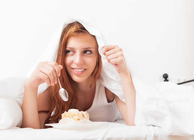 Junge Frau, die süßen Kuchen unter Blatt im Bett isst