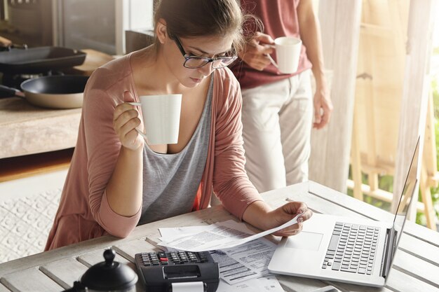 Junge Frau, die Tee trinkt und Rechnung in ihren Händen studiert, frustrierten Blick, während sie Familienbudget verwaltet und Papierkram tut, am Küchentisch mit Papieren, Taschenrechner und Laptop-Computer sitzt