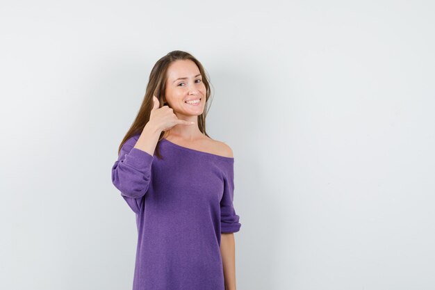 Junge Frau, die Telefongeste im violetten Hemd zeigt und hilfreich schaut. Vorderansicht.