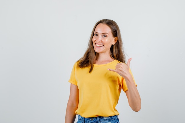 Junge Frau, die Telefongeste in T-Shirt, Shorts zeigt und fröhlich schaut