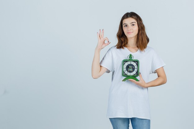 Junge Frau, die Uhr hält, zeigt ok Geste im weißen T-Shirt, Jeans und schaut fröhlich, Vorderansicht.