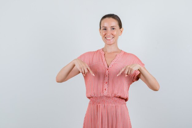 Junge Frau, die unten im gestreiften Kleid zeigt und fröhlich, Vorderansicht schaut.