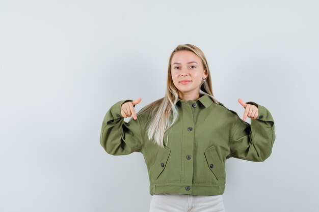 Junge Frau, die unten in der grünen Jacke, in den Jeans zeigt und glücklich schaut. Vorderansicht.