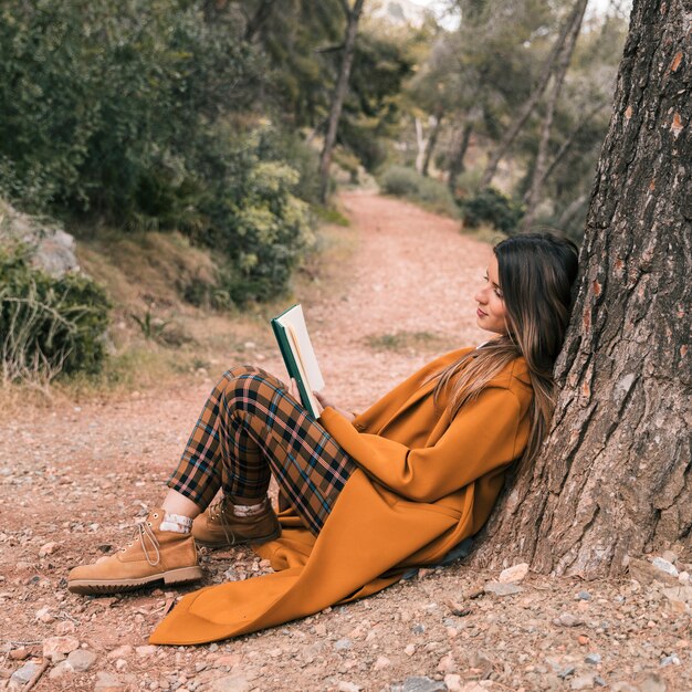 Junge Frau, die unterwegs unter dem Baum Lesebuch sitzt
