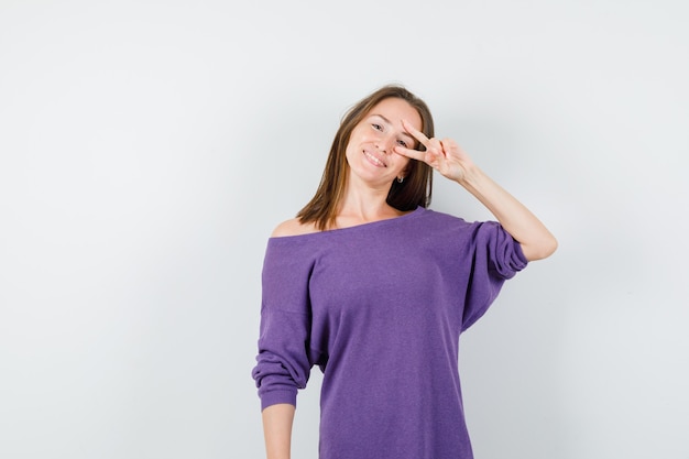 Junge Frau, die V-Zeichen auf Auge im violetten Hemd zeigt und fröhlich schaut. Vorderansicht.