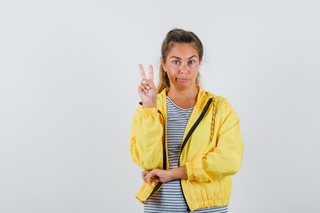 Junge Frau, die V-Zeichen im T-Shirt, in der Jacke zeigt und nachdenklich, Vorderansicht schaut.