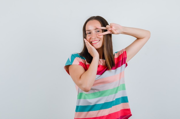 Junge Frau, die V-Zeichen nahe Auge im T-Shirt zeigt und glückliche Vorderansicht schaut.