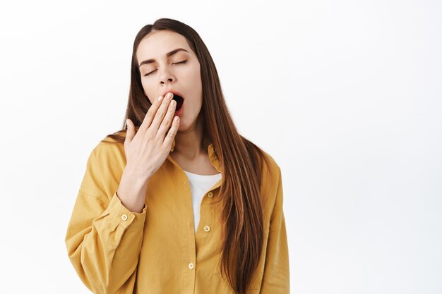 Junge Frau, die vor Vergnügen gähnt, den offenen Mund mit der Handfläche bedecken und schläfrigen Augen geschlossen stehen, früh morgens aufwachen, Kaffee brauchen, weiße Wand