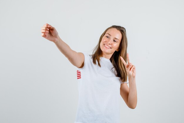 Junge Frau, die während des Siegeszeichens im weißen T-Shirt aufwirft und glücklich schaut. Vorderansicht.