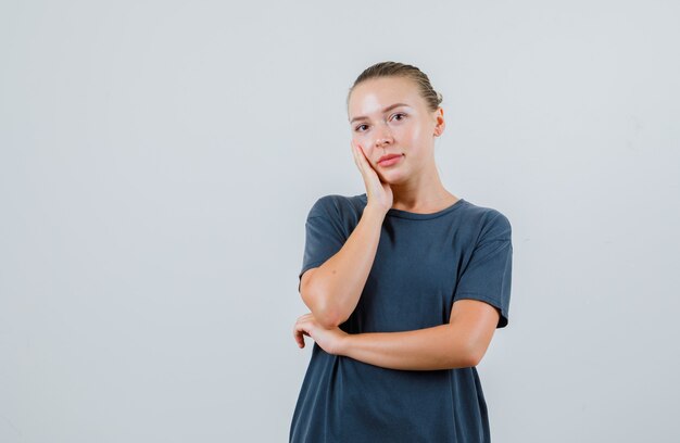 Junge Frau, die Wange auf erhabener Handfläche im grauen T-Shirt lehnt und nachdenklich schaut