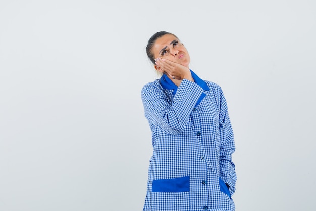 Junge Frau, die Wange auf Handfläche lehnt, in denkender Geste im blauen Gingham-Pyjamahemd stehend und nachdenklich, Vorderansicht schauend steht.