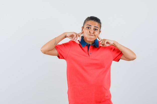 Junge Frau, die Wangen aufbläst, Zeigefinger auf Wangen im roten T-Shirt setzt und hübsch aussieht, Vorderansicht.