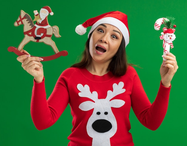 Kostenloses Foto junge frau, die weihnachtsweihnachtsmütze und roten pullover trägt, der weihnachtsspielzeug hält, das die kamera lächelt, die fröhlich über grünem hintergrund steht