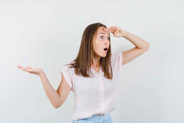 Junge Frau, die weit weg mit ausgebreiteter Handfläche im T-Shirt, im Rock schaut und erstaunt schaut. Vorderansicht.