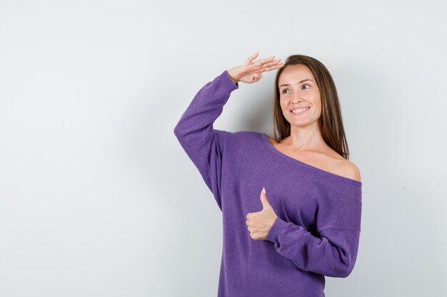 Junge Frau, die weit weg mit Daumen oben im violetten Hemd schaut und optimistisch, Vorderansicht schaut.