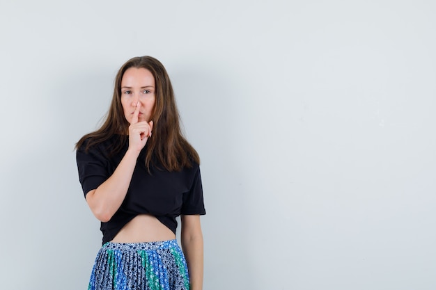 Junge Frau, die Zeigefinger auf Mund setzt und Stille Geste in schwarzem T-Shirt und blauem Rock zeigt und attraktiv aussieht