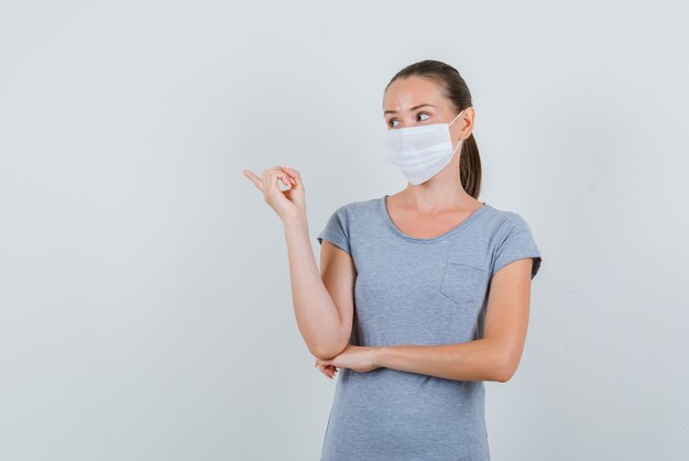 Junge Frau, die zur Seite im grauen T-Shirt zeigt, Maskenvorderansicht.