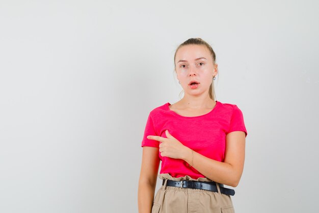 Junge Frau, die zur Seite im T-Shirt, in der Hose zeigt und überrascht sieht, Vorderansicht.