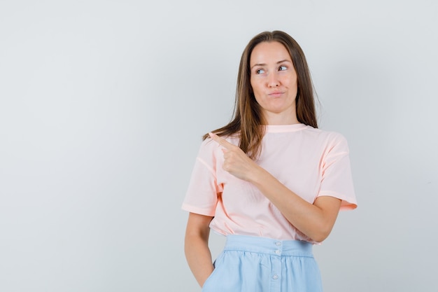 Junge Frau, die zur Seite in T-Shirt, Rock zeigt und gerissen aussieht. Vorderansicht.