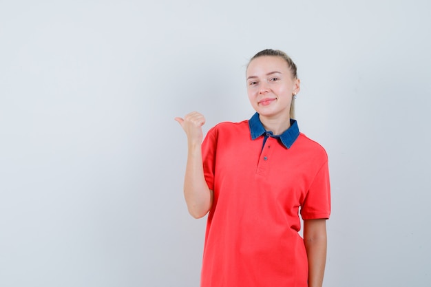 Junge Frau, die zur Seite mit Daumen im T-Shirt zeigt und lustig schaut